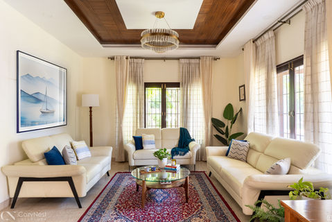Bright, transitional living room featuring three beige sofas, blue and white throw pillows, a glass coffee table over a patterned red and blue rug, a large sailboat painting, and a rectangular tray ceiling with warm wood paneling and a crystal chandelier.
