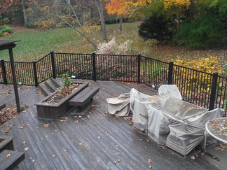Wooden deck with black railing, covered furniture, and fall foliage in the background.