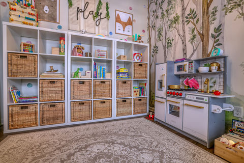 Children’s playroom with white cubby storage and wicker baskets, play kitchen set, and tree-patterned wallpaper for a woodland theme.