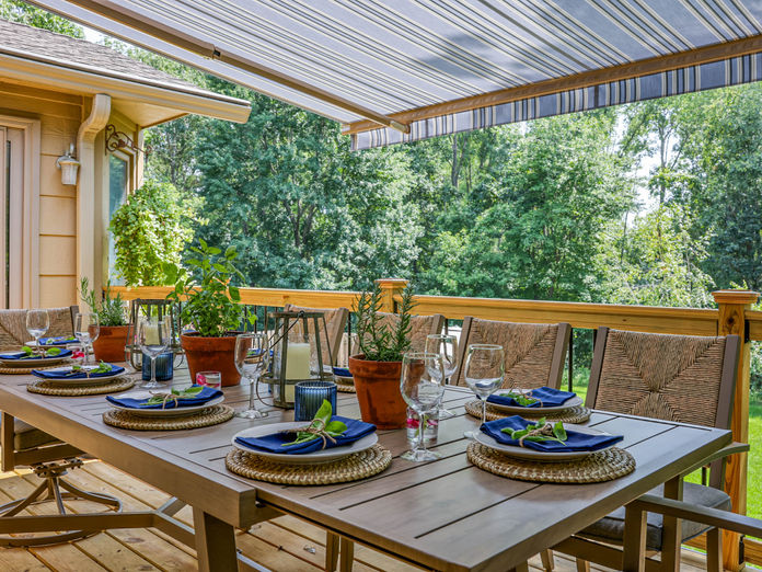 Garden Dining transformation with a table set for six, layered with woven placemats, blue napkins, and potted herbs. A new canopy provides shade while the greenery creates a relaxed outdoor retreat.