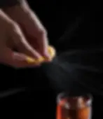Hände pressen eine Orangenschale über einem Cocktail aus