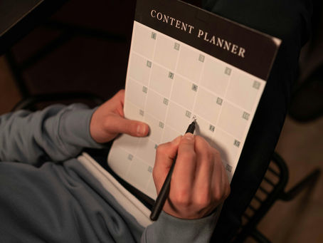 A person with a light blue long-sleeved shirt is seen from an overhead view, holding a black 'Content Planner' calendar. Their right hand is using a black pen to write on or mark a date, possibly the 12th or 13th, on the white grid. The calendar is titled 'CONTENT PLANNER' in white text on a black bar.