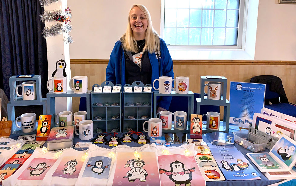 Smiling woman in a blue jacket stands behind a table displaying penguin-themed mugs and art. A festive atmosphere with lights and decorations.