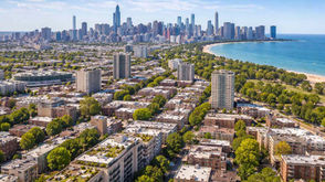 Aerial view of Chicago’s Lakeview neighborhood featuring mid rise residential buildings, tree lined streets, Lake Michigan shoreline, and the downtown skyline in the background on a clear sunny day.