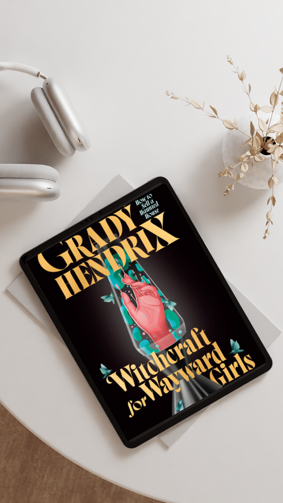 Tablet displaying "Grady Hendrix: Witchcraft for Wayward Girls" cover on a table with headphones and vase. Black background, bold text.