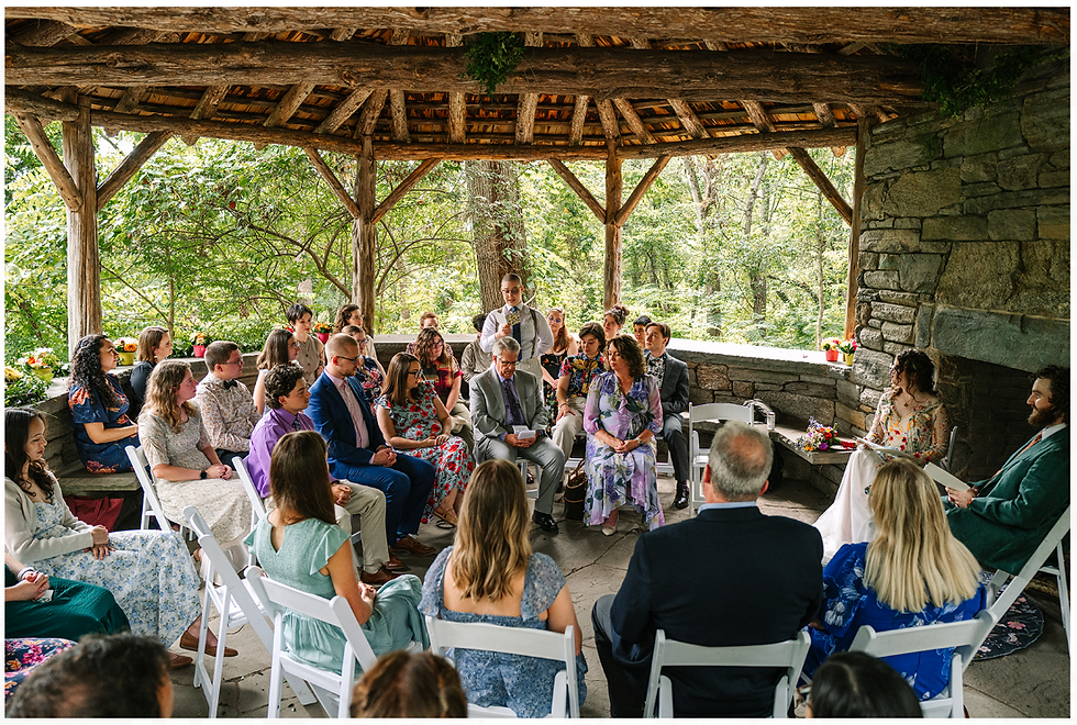 Guests sit in a rustic open pavilion during a wedding ceremony, with greenery and stone walls behind them.
