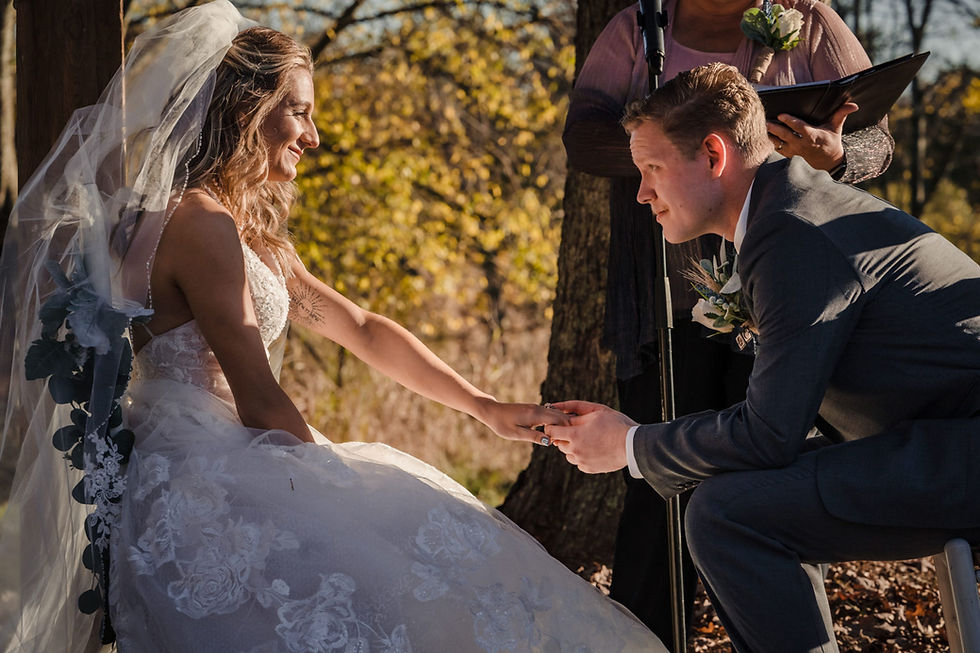 Bride and groom hold hands at an outdoor wedding in a peaceful rural setting, while the officiant stands behind them reading from a decorative binder.