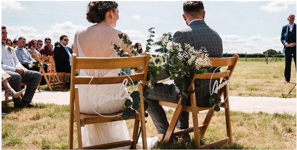 Bride and groom sit on decorated wooden chairs at an outdoor wedding, facing guests and officiant under a bright sky.