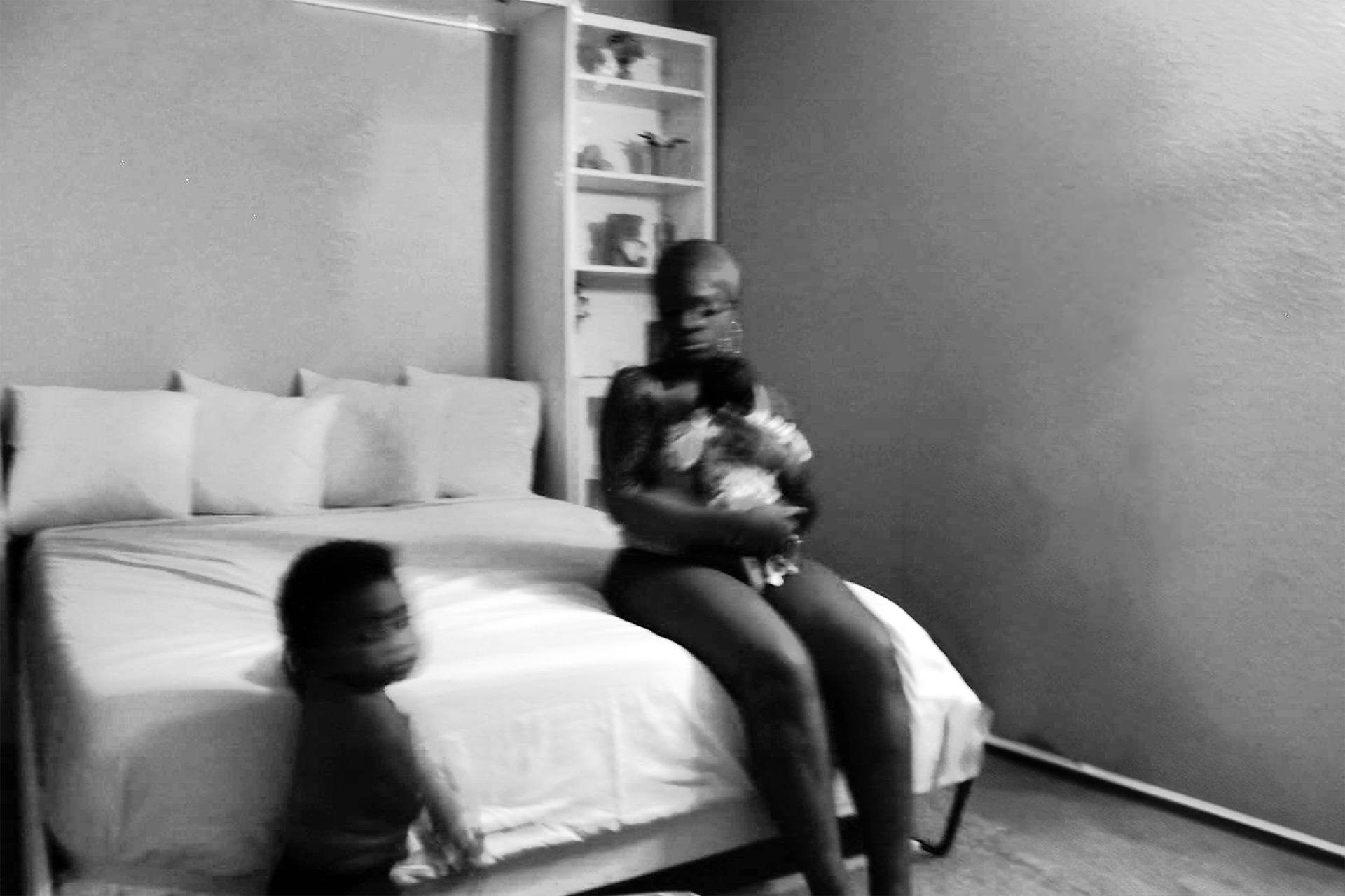 woman on a bed with 2 kids one in hand the other free to roam at the foot of the bed
