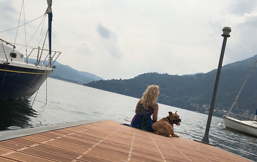 Woman with dog sitting on dock, overlooking lake; sailboats in background.