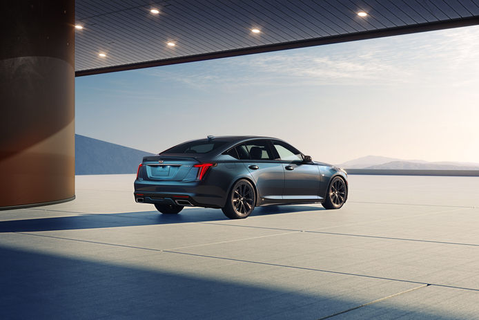 A blue Cadillac sedan parked under a sunlit canopy with several spotlights, facing a landscape and casting a long shadow