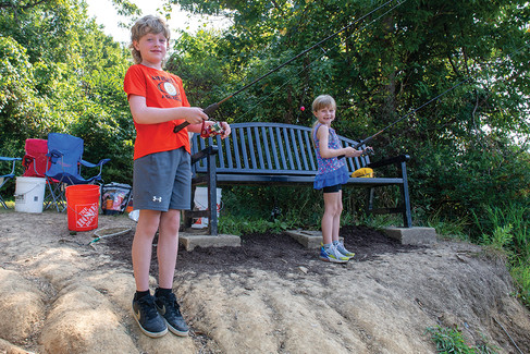 A boy with light hair and an orange t-shirt and a girl with blonde pigtails and a purple top smile at the camera while holding fishing poles. They are standing on a dirt bank next to a black park bench, with fishing gear scattered nearby.