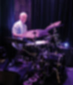A candid photograph of a senior Caucasian man with short, grey hair, looking directly at the camera while sitting behind a multi-cymbal black drum kit on a stage. He wears a light-blue button-down shirt. The scene is bathed in cool purple and blue stage lighting, which illuminates detailed black Zildjian cymbal cases to his left, intricate metallic Art Deco-style panels behind him, and the edge of a stage monitor in the foreground.