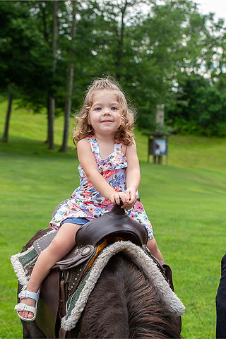 A young girl with curly hair smiles while riding a pony at a festival.