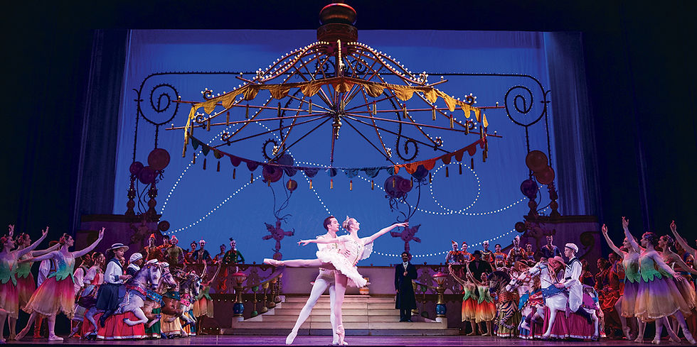 A lavish ballet production of The Nutcracker on stage, featuring the Sugar Plum Fairy and her Cavalier performing a pas de deux center stage. They are surrounded by a large ensemble cast, elaborate period costumes, and a magnificent, carousel-like set piece with hanging decorations.