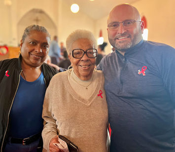Mayor Tom Maglicco (right), wearing glasses and a grey Under Armour quarter-zip pullover, stands with his arm around a smiling elderly woman in a beige v-neck sweater. A third woman in a blue shirt and black jacket stands to the left. All three are wearing pink breast cancer awareness ribbons pinned to their tops. They are gathered inside a brightly lit community space with a vaulted ceiling.