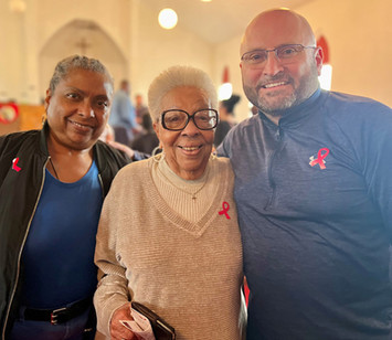 Mayor Tom Maglicco (right), wearing glasses and a grey Under Armour quarter-zip pullover, stands with his arm around a smiling elderly woman in a beige v-neck sweater. A third woman in a blue shirt and black jacket stands to the left. All three are wearing pink breast cancer awareness ribbons pinned to their tops. They are gathered inside a brightly lit community space with a vaulted ceiling.
