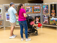 A family of three views an outdoor art exhibit display. A man in a light blue shirt and a woman in a magenta shirt with a ponytailed child in a black stroller are looking closely at various framed prints and canvases mounted on metal grid panels. The art includes landscape photography, colorful cityscapes, and a pop-art style portrait. The ground is paved with large light-colored tiles.