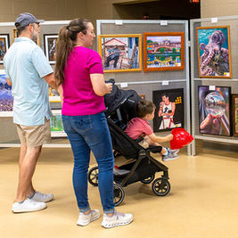 A family of three views an outdoor art exhibit display. A man in a light blue shirt and a woman in a magenta shirt with a ponytailed child in a black stroller are looking closely at various framed prints and canvases mounted on metal grid panels. The art includes landscape photography, colorful cityscapes, and a pop-art style portrait. The ground is paved with large light-colored tiles.