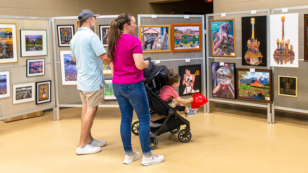 A family of three views an outdoor art exhibit display. A man in a light blue shirt and a woman in a magenta shirt with a ponytailed child in a black stroller are looking closely at various framed prints and canvases mounted on metal grid panels. The art includes landscape photography, colorful cityscapes, and a pop-art style portrait. The ground is paved with large light-colored tiles.