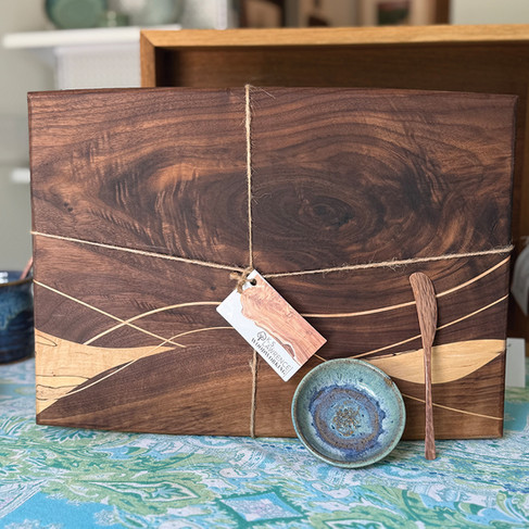 A large, beautifully crafted walnut wood charcuterie board with a lighter-colored maple or ash inlay in a flowing wave pattern. It is wrapped with twine and features a small, ceramic "handmade" tag. A miniature blue and purple glazed ceramic dish and a small wooden butter knife are placed near the tag, all resting on a blue and white patterned paisley table runner.
