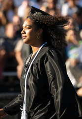 A side-profile view of a female graduate with voluminous, dark curly hair walking outdoors in the sunshine. She is wearing a black graduation gown, hoop earrings, and a white honor cord draped over her shoulder. The background features a softly blurred crowd of spectators in stadium seating.
