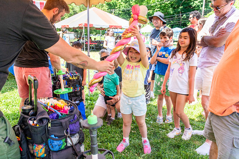 A close-up photograph of a balloon artist interacting with children at a festival. From the left, a man with a tattooed forearm and a black shirt extends a newly made, twisted pink and gold balloon sculpture towards a joyful young girl. The girl, wearing a yellow shirt, patterned shorts, a pink sun hat, and pink shoes, reaches up with both hands to receive the balloon with an ecstatic expression. Other children and adults watch from behind a rope barrier. The artist's elaborate backpack full of balloons and tools is visible in the foreground.