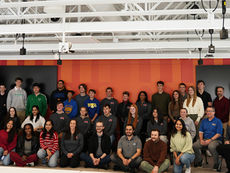 A wide-angle group portrait of approximately 35 students and adults associated with the Human-Centered AI project, posing in a modern room with exposed ceiling beams. The group is arranged in three rows—some sitting on the floor, some in chairs, and others standing—against a vibrant orange and red paneled wall with two large black screens.  The students are wearing various school-affiliated clothing, including the matching grey West Allegheny "WA" quarter-zips and hoodies representing the Cornell, Peters Township, and South Fayette school districts. Faculty advisors and CMU representatives are interspersed throughout the group, capturing a collaborative regional partnership between the four school districts and Carnegie Mellon University.