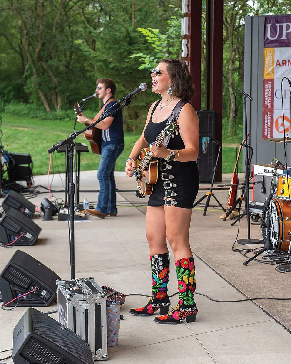 A female musician in a black dress and colorful cowboy boots sings and plays bass guitar on an outdoor stage, with a male guitarist in the background.