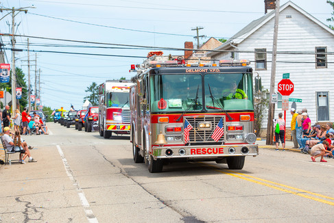 A Delmont V.F.D. fire rescue truck leading a line of emergency and parade vehicles down a street lined with spectators. The red and silver fire truck has 'DELMONT V.F.D.' and 'RESCUE' clearly visible.