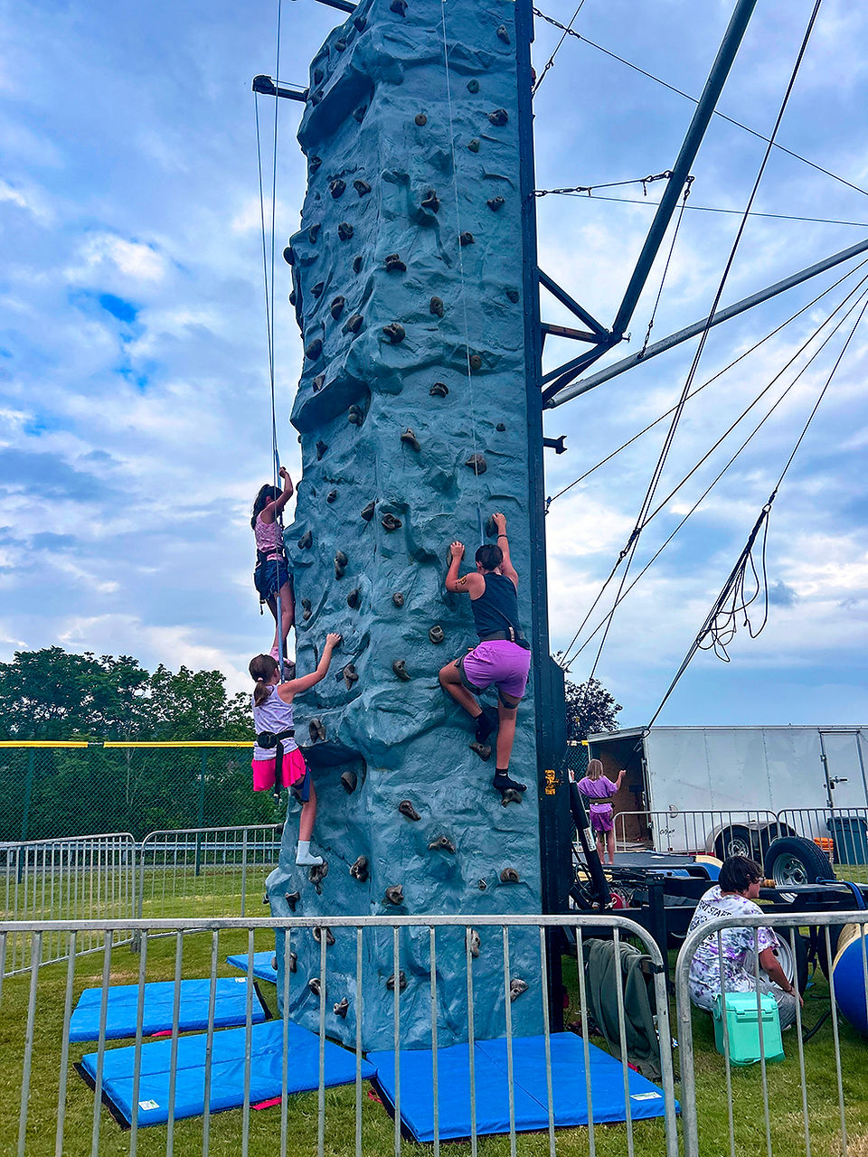 A medium shot of a portable, trailer-mounted rock climbing wall on a grassy area under a cloudy sky. Three children are actively climbing the textured blue wall. The two girls on the left are near the bottom, one wearing a white and pink top and a pink skirt, the other in a purple tank top and purple shorts. A boy is higher up on the right, also climbing. All wear harnesses and are connected to belay ropes. Blue crash pads are at the base of the wall. Metal crowd control barriers separate the climbing wall from the foreground. In the background are a white utility trailer, a few trees, and the edge of a sports field fence. A person in a tie-dye shirt sits near a cooler to the right.