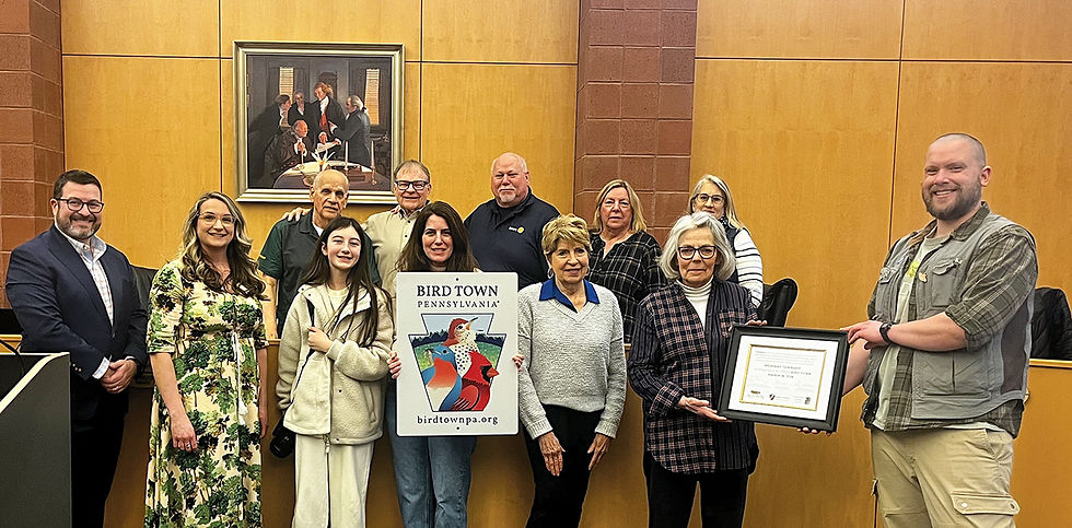 A medium wide shot in color shows 11 people in casual clothing smiling in front of wood paneled walls in a meeting room, holding a framed certificate and a rectangular sign with a Keystone shaped logo in the center and the words "BIRD TOWN PENNSYLVANIA birdtownpa.org" below the logo. At the top of the sign in large black capital letters are the words "BIRD TOWN PENNSYLVANIA". In the center of the sign is a blue and red Keystone shape with a drawing of a red and blue and yellow bird perched in the center with a patch of blue sky and clouds in the upper right. Directly below the Keystone logo in small red capital letters are the words "birdtownpa.org". Starting from the far left, a man in a black sportcoat and glasses stands with his hands in front, a woman in a floral dress stands with her arms down, an older man in a green t-shirt stands behind a young girl in a cream jacket, next to him a bald older man in a red polo shirt, and in front an older woman with blonde hair wearing a grey sweater, next to her an older woman with white hair wearing a plaid button down shirt, next to her a man with a short beard in a green button down vest holds the framed certificate. Standing behind the older woman wearing the grey sweater is an older woman with blonde hair, wearing a grey and black sweater, next to her another woman with blonde hair and glasses. On the left in the background on the wall is a rectangular gold framed painting of five men in business attire seated and standing around a table with a white tablecloth. The group is centered in the frame.