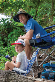 A man wearing a patriotic-themed bucket hat and sunglasses sits in a blue camping chair, smiling at the camera. In front of him, a young boy in a baseball cap and a white t-shirt sits in a smaller blue chair, holding a fishing pole and also smiling at the camera. They are on a shaded, wooded bank by a pond.