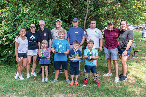 A large group of adults and children stands in a grassy area in front of a green, leafy background. On the left, an adult woman in a white t-shirt and shorts stands next to an adult woman in a black "MAKE AN IMPACT" t-shirt and white shorts. A young girl with a trophy stands in front of them. In the center, a man in a black polo shirt and a man in a tan polo shirt stand behind a young boy in a light blue t-shirt and dark shorts, who holds a trophy and certificate. Next to them, a man in a blue t-shirt stands behind a younger boy in a dark blue shirt and shorts, who also holds a trophy. On the right, a man in a white t-shirt stands next to a man in a dark red polo shirt and shorts. A young boy in a light blue t-shirt, shorts, and a baseball cap holds a trophy and certificate in front of them. On the far right, an adult woman in a black t-shirt and jean shorts stands next to the man in the red polo. All the children are smiling and holding trophies.