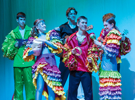A group of performers on stage wear vibrant, colorful costumes with layered, rainbow-colored ruffles, singing and holding musical instruments like maracas and a tambourine under soft blue lighting.
