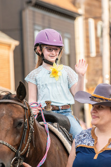 A smiling girl in a purple helmet, glasses, and a blue shirt with a yellow flower is seated on a brown horse during the parade.