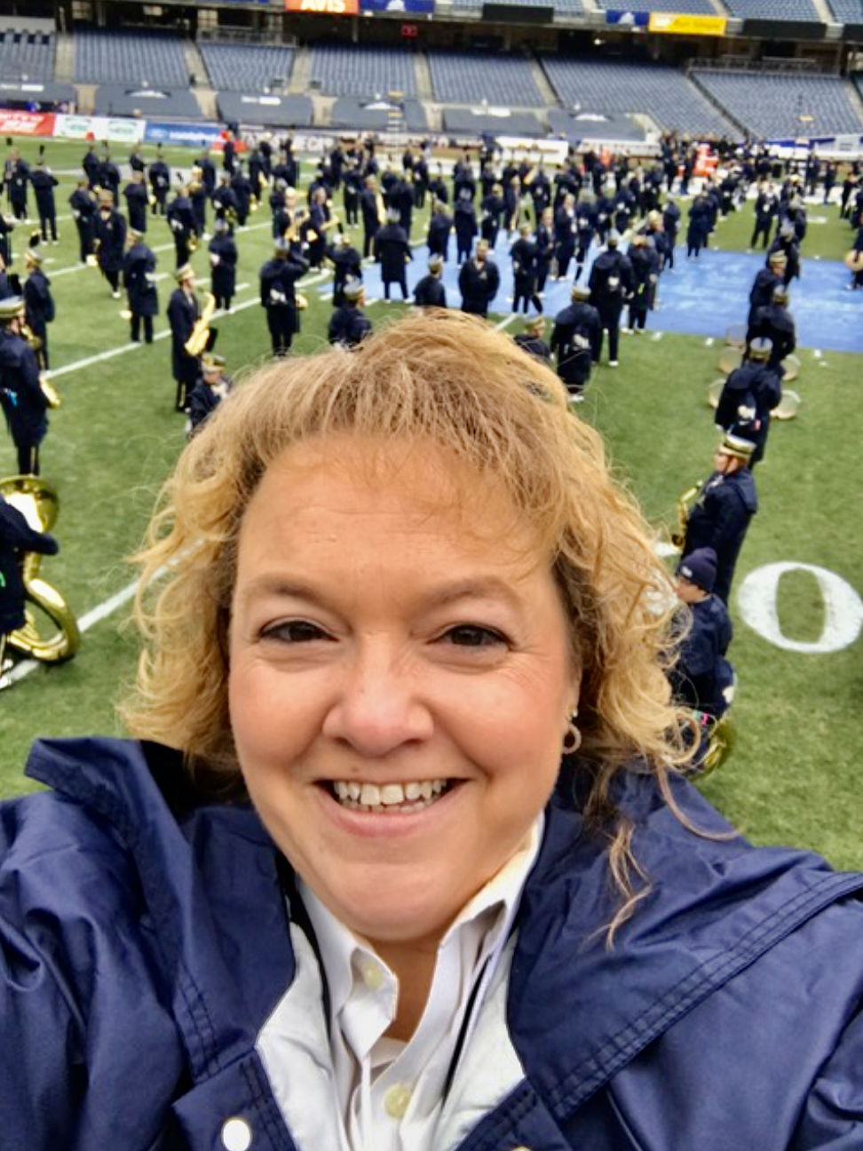 Selfie of Virginia Schaap smiling with a marching band practicing on a football field in the background.