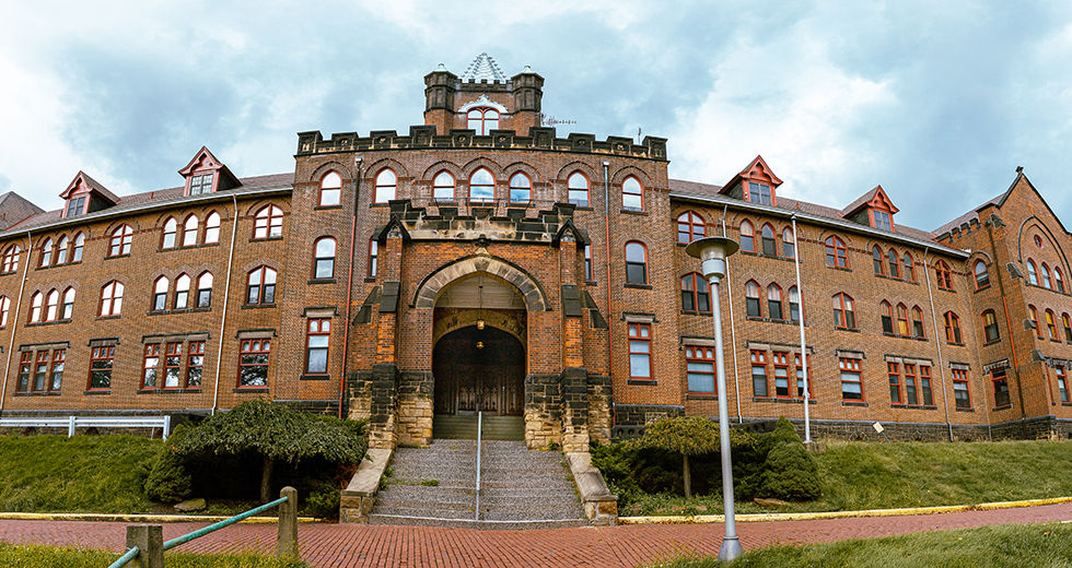 A wide-angle, panoramic photograph of the historic Mt. Alvernia Motherhouse. The large, three-story brick building features distinctive Gothic Revival architecture with arched windows, red trim, and a prominent central stone entrance with a large arched doorway. A central tower with a decorative peaked roof sits atop the building against a cloudy sky. A brick walkway and a green grassy slope lead up to the structure.