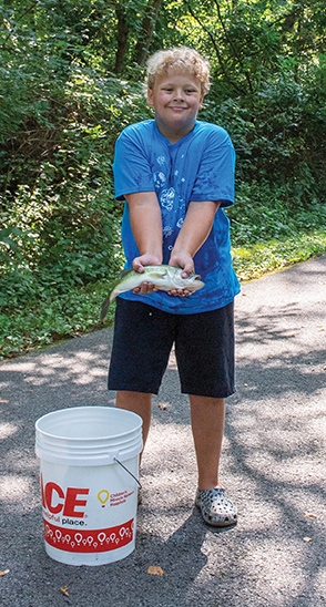 A young boy with blond hair and a blue t-shirt stands on a paved path, holding a fish in his cupped hands. A white bucket with a "Miller's Ace Hardware" logo is beside him.