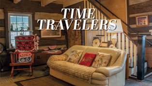 The rustic interior living room of the Wyckoff-Mason House features a cream-colored couch with a cozy throw and decorative pillows. The room has exposed wooden walls, a rocking chair with a "Route 66" pillow, and a wooden staircase leading upstairs.
