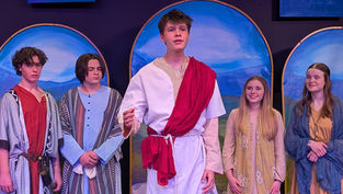 Jude Glover, dressed in a white tunic with a red sash as Jesus, stands center stage singing during the production of "Another Miracle." To his right, Sylvia Youshack as Mary and another actress look on with emotional expressions, while Leo Pietroforte and another male performer stand to the left in patterned biblical robes. They are performing in front of three arched backdrops painted with blue skies and mountains.