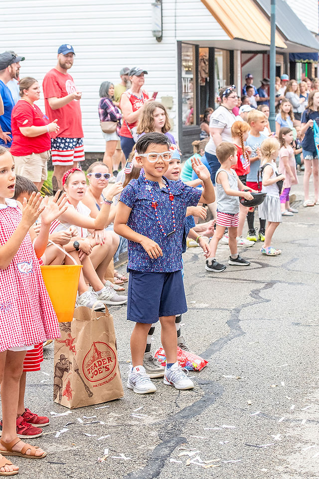 A young boy wearing festive glasses and star-spangled beads standing on a street curb among a crowd of spectators, smiling and waving during a town parade.