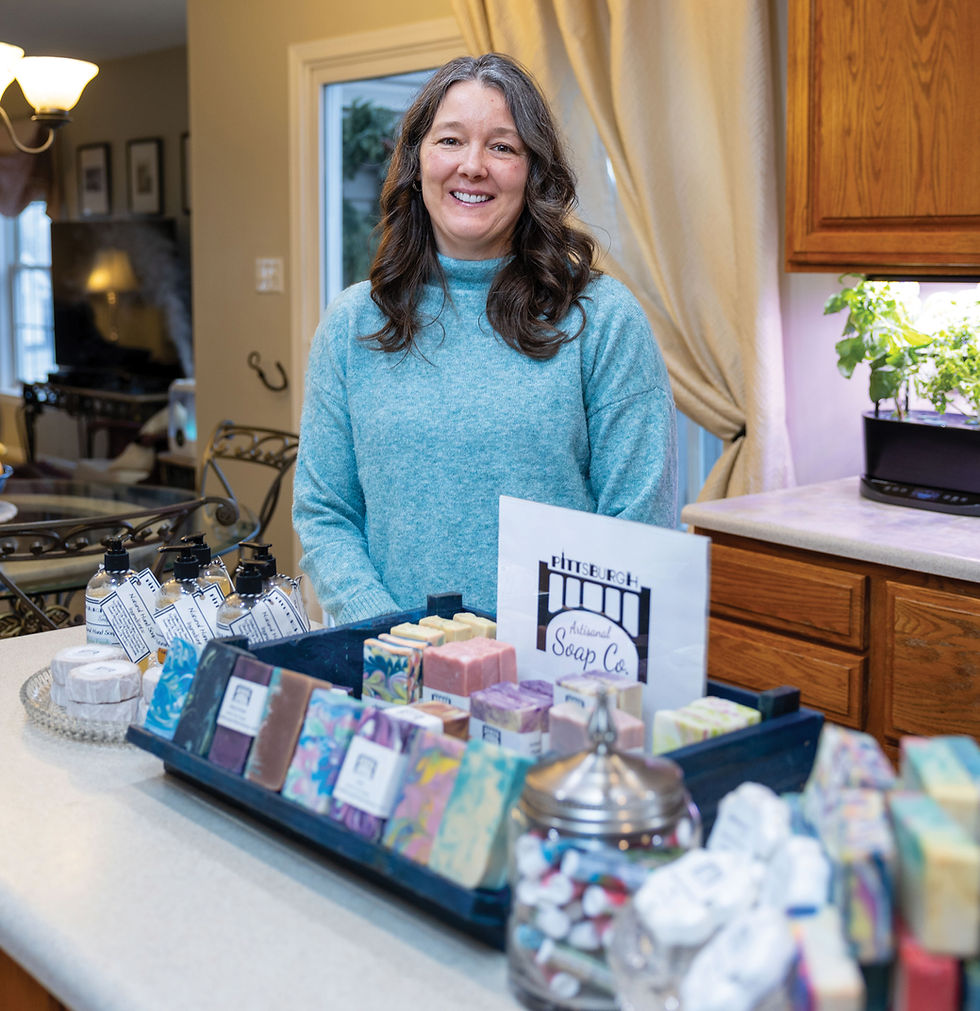 Laura Bogacz, founder of Pittsburgh Artisanal Soap Co., smiles behind a display of her colorful, handmade bar soaps. She is wearing a blue sweater, and the foreground features a wooden tray filled with patterned soap bars and a glass jar of small soap samples.