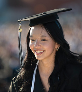 A bright, close-up portrait of a smiling female graduate of East Asian descent looking to the left. She wears a black graduation cap with a black and white tassel featuring a gold "26" year charm. White honor cords are draped over her shoulders, and a blurred crowd fills the warm, sunlit background.