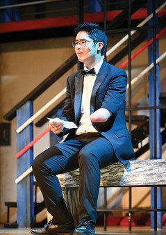 A student performs a reflective moment in Greensburg Salem High School’s spring musical Anything Goes, seated on a stage bench in a black tuxedo and bow tie while holding a note in one hand. Warm and cool stage lighting falls across the ship-themed set and staircase behind him, emphasizing the quiet, thoughtful tone of the scene.