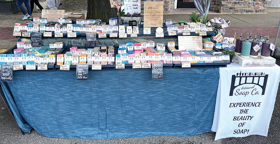 A detailed outdoor market display for Pittsburgh Artisanal Soap Co. featuring rows of colorful, handmade soap bars arranged on tiered shelving. The table is draped in a blue cloth, with clear signage indicating various scents like "Ocean," "Fruity," and "Floral," alongside a white banner that reads, "Experience the Beauty of Soap!"