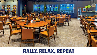 The spacious interior dining area of Baseline Bar & Grille, featuring numerous wooden tables and tan chairs set for service. Large windows in the background provide a clear view of blue-lit indoor pickleball courts, while decorative dartboards with chevron patterns line the left wall.