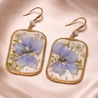 A pair of rectangular, gold-rimmed drop earrings featuring pressed purple flowers and small decorative accents encased in clear resin, displayed on a soft, light-colored satin fabric.