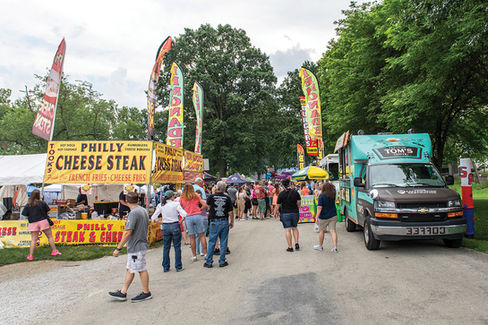 A line of people walking toward food trucks and vendor tents, with large banner signs advertising "PHILLY CHEESE STEAK" and other items.