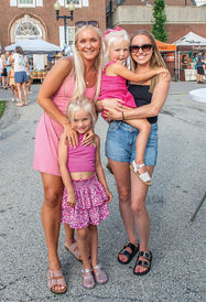 A group of four smiling females—two women and two young girls—are posed together for a photo on an asphalt street during a daytime outdoor market. The woman on the left, with long blonde hair and wearing a pink sun dress, has her arm around the younger girl in front of her, who wears a pink ribbed crop top and a purple floral skirt. The woman on the right, wearing a black tank top, denim shorts, and sunglasses, holds the older girl on her hip, who wears a pink tiered dress. All are looking at the camera. In the background are market tents, brick buildings, and other attendees.